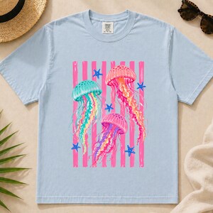 K&ouml;nnte beinhalten: Ein hellblaues kurz&auml;rmeliges T-Shirt mit einem Grafikdruck von drei Quallen in Rosa-, Gelb- und Blaugr&uuml;nt&ouml;nen. Das Design enth&auml;lt vertikale rosa Streifen und kleine blaue Seestern-Akzente. Das T-Shirt hat einen Rundhalsausschnitt und ein Etikett.