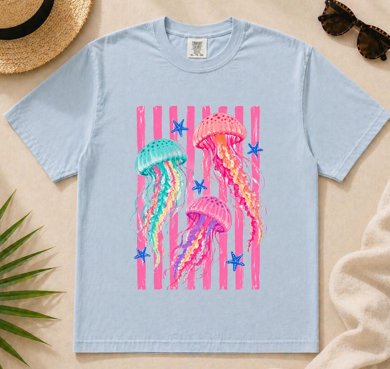 K&ouml;nnte beinhalten: Ein hellblaues kurz&auml;rmeliges T-Shirt mit einem Grafikdruck von drei Quallen in Rosa-, Gelb- und Blaugr&uuml;nt&ouml;nen. Das Design enth&auml;lt vertikale rosa Streifen und kleine blaue Seestern-Akzente. Das T-Shirt hat einen Rundhalsausschnitt und ein Etikett.