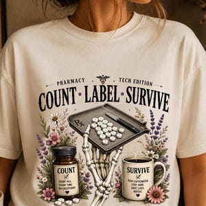 May include: Cream-colored t-shirt with a pharmacy-themed graphic. The design includes the words "COUNT LABEL SURVIVE" with a pill tray, a skeleton hand, a coffee mug, and a pill bottle. Floral elements and text are also present.