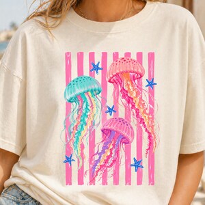 K&ouml;nnte beinhalten: Ein cremefarbenes Kurzarm-T-Shirt mit einem Grafikdruck von drei Quallen in T&uuml;rkis-, Rosa- und Violettt&ouml;nen. Das Design zeigt vertikale rosa Streifen und kleine blaue Seesterne, die &uuml;ber die Vorderseite des T-Shirts verteilt sind.
