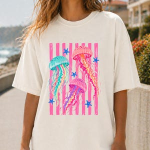K&ouml;nnte beinhalten: Ein cremefarbenes Kurzarm-T-Shirt mit einem Grafikdruck von drei Quallen in T&uuml;rkis-, Pink- und Violettt&ouml;nen. Das Design zeigt vertikale pinke Streifen und kleine blaue Seesterne, die mittig auf der Vorderseite des Kleidungsst&uuml;cks platziert sind.