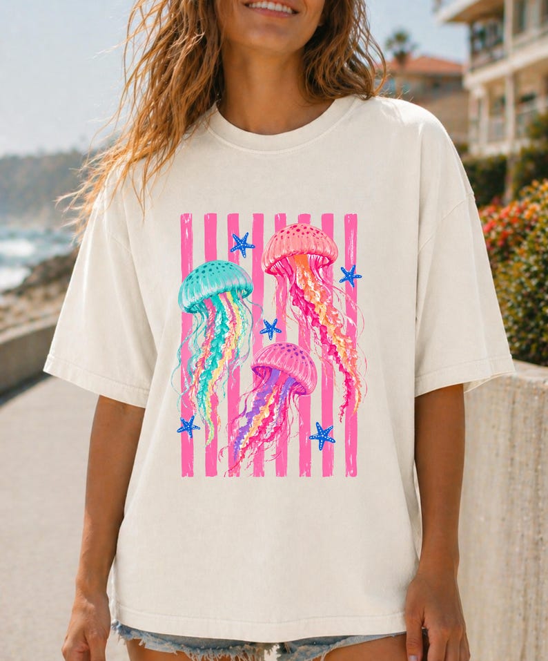 K&ouml;nnte beinhalten: Ein cremefarbenes Kurzarm-T-Shirt mit einem Grafikdruck von drei Quallen in T&uuml;rkis-, Pink- und Violettt&ouml;nen. Das Design zeigt vertikale pinke Streifen und kleine blaue Seesterne, die mittig auf der Vorderseite des Kleidungsst&uuml;cks platziert sind.