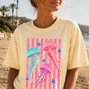 K&ouml;nnte beinhalten: Ein hellgelbes Kurzarm-T-Shirt mit einem Grafikdruck von drei Quallen in T&uuml;rkis-, Rosa- und Violettt&ouml;nen. Das Design zeigt vertikale rosa Streifen und kleine blaue Seesterne, die &uuml;ber die Vorderseite des Kleidungsst&uuml;cks verteilt sind.