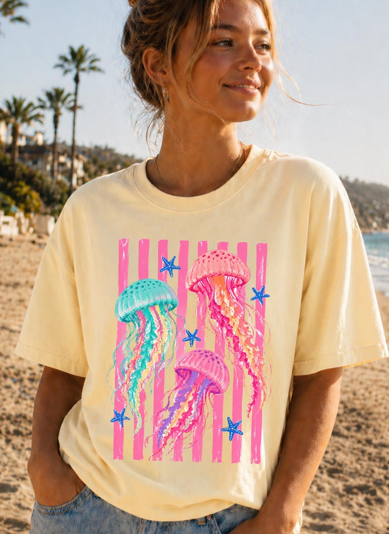 K&ouml;nnte beinhalten: Ein hellgelbes Kurzarm-T-Shirt mit einem Grafikdruck von drei Quallen in T&uuml;rkis-, Rosa- und Violettt&ouml;nen. Das Design zeigt vertikale rosa Streifen und kleine blaue Seesterne, die &uuml;ber die Vorderseite des Kleidungsst&uuml;cks verteilt sind.