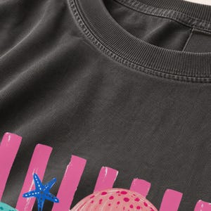 K&ouml;nnte beinhalten: Ein anthrazitfarbenes T-Shirt mit einem Grafikdruck aus vertikalen rosa Streifen, einem blauen Seestern und einer rosa gepunkteten Muschel. Das Shirt hat einen gerippten Rundhalsausschnitt und eine verwaschene Stofftextur auf der gesamten Oberfl&auml;che.