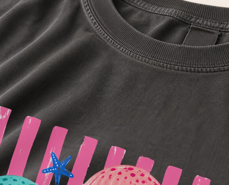 K&ouml;nnte beinhalten: Ein anthrazitfarbenes T-Shirt mit einem Grafikdruck aus vertikalen rosa Streifen, einem blauen Seestern und einer rosa gepunkteten Muschel. Das Shirt hat einen gerippten Rundhalsausschnitt und eine verwaschene Stofftextur auf der gesamten Oberfl&auml;che.