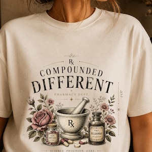 May include: Cream-colored t-shirt featuring a vintage pharmacy design. The graphic includes a mortar and pestle, bottles, flowers, and the text "COMPOUNDED DIFFERENT PHARMACY DEPT." in a decorative font. The design has a botanical and apothecary theme.