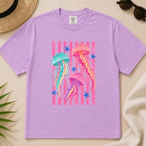 K&ouml;nnte beinhalten: Ein hellviolettes Kurzarm-T-Shirt mit einem Grafikdruck von drei bunten Quallen in T&uuml;rkis-, Rosa- und Oranget&ouml;nen, umgeben von kleinen blauen Seesternen vor einem Hintergrund aus vertikalen rosa Streifen auf der Brustmitte.