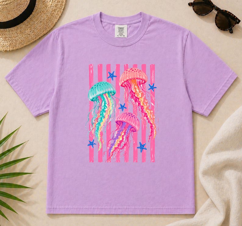 K&ouml;nnte beinhalten: Ein hellviolettes Kurzarm-T-Shirt mit einem Grafikdruck von drei bunten Quallen in T&uuml;rkis-, Rosa- und Oranget&ouml;nen, umgeben von kleinen blauen Seesternen vor einem Hintergrund aus vertikalen rosa Streifen auf der Brustmitte.