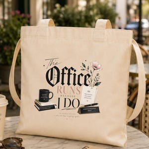 以下が含まれることがあります： The Office Runs Because I Doという黒とピンクのグラフィックプリントが施されたベージュのキャンバストートバッグ。デザインには、黒いコーヒーマグ、積み重ねられた本、ペン、デスクネームプレート、小さなピンクの花のイラストが含まれています。