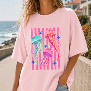 K&ouml;nnte beinhalten: Ein hellrosa T-Shirt mit kurzen &Auml;rmeln und einem zentralen Aufdruck von drei bunten Quallen in T&uuml;rkis, Orange und Violett, umgeben von kleinen blauen Seesternen vor einem Hintergrund aus vertikalen rosa Streifen.