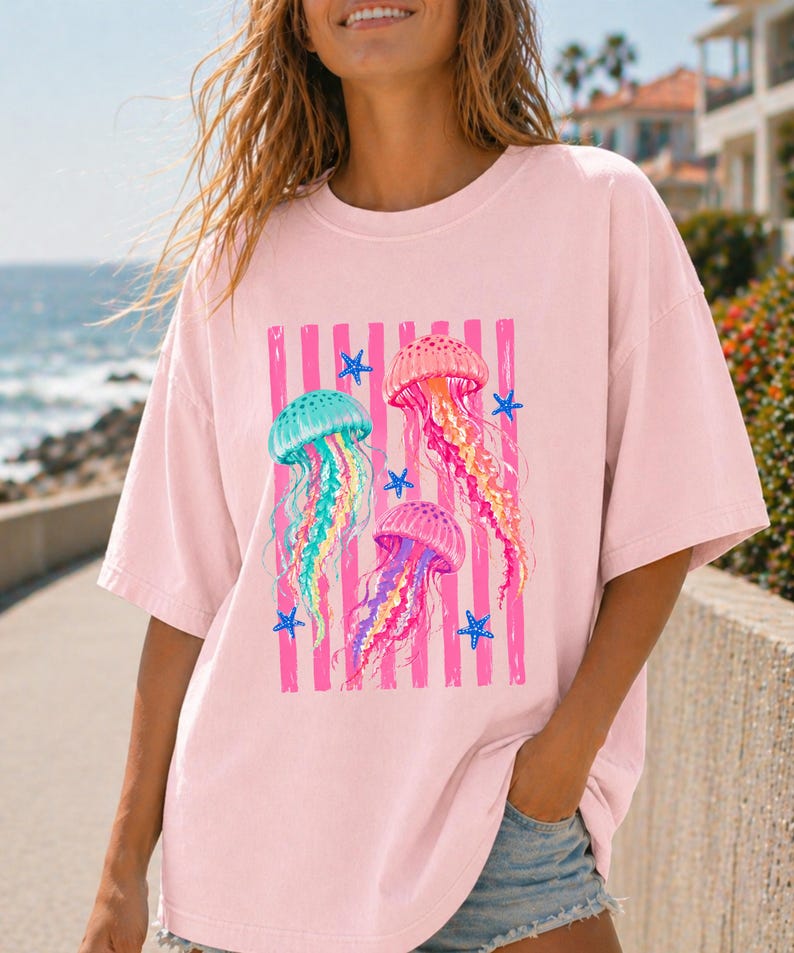 K&ouml;nnte beinhalten: Ein hellrosa T-Shirt mit kurzen &Auml;rmeln und einem zentralen Aufdruck von drei bunten Quallen in T&uuml;rkis, Orange und Violett, umgeben von kleinen blauen Seesternen vor einem Hintergrund aus vertikalen rosa Streifen.