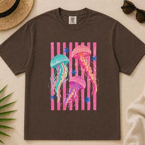 K&ouml;nnte beinhalten: Ein dunkelbraunes Kurzarm-T-Shirt mit einem Grafikdruck von drei Quallen in Rosa-, Gelb- und T&uuml;rkist&ouml;nen. Das Design zeigt vertikale rosa Streifen und kleine blaue Seestern-Akzente. Das T-Shirt hat einen Rundhalsausschnitt und ein Etikett.