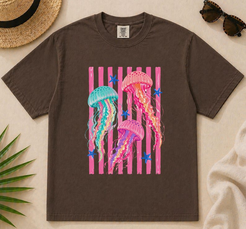 K&ouml;nnte beinhalten: Ein dunkelbraunes Kurzarm-T-Shirt mit einem Grafikdruck von drei Quallen in Rosa-, Gelb- und T&uuml;rkist&ouml;nen. Das Design zeigt vertikale rosa Streifen und kleine blaue Seestern-Akzente. Das T-Shirt hat einen Rundhalsausschnitt und ein Etikett.