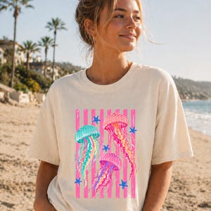 K&ouml;nnte beinhalten: Ein cremefarbenes Kurzarm-T-Shirt mit einem Grafikdruck von drei Quallen in Rosa-, Blau- und Gelbt&ouml;nen, akzentuiert durch kleine blaue Seesterne und vertikale rosa Streifen auf der Vorderseite des Kleidungsst&uuml;cks.