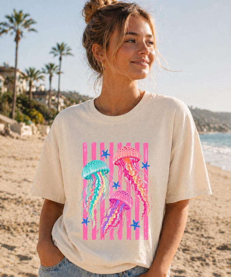 K&ouml;nnte beinhalten: Ein cremefarbenes Kurzarm-T-Shirt mit einem Grafikdruck von drei Quallen in Rosa-, Blau- und Gelbt&ouml;nen, akzentuiert durch kleine blaue Seesterne und vertikale rosa Streifen auf der Vorderseite des Kleidungsst&uuml;cks.