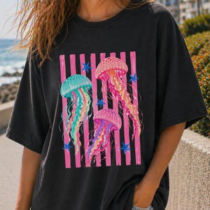 K&ouml;nnte beinhalten: Ein schwarzes Kurzarm-T-Shirt mit einem Grafikdruck von drei Quallen in den Farben Pink, T&uuml;rkis und Gelb. Das Design zeigt vertikale pinke Streifen hinter den Quallen sowie mehrere kleine blaue Seesterne, die um das zentrale Motiv verteilt sind.