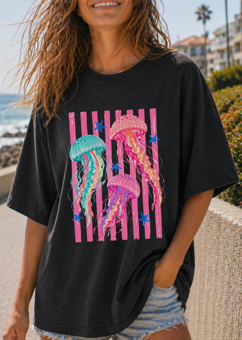 K&ouml;nnte beinhalten: Ein schwarzes Kurzarm-T-Shirt mit einem Grafikdruck von drei Quallen in den Farben Pink, T&uuml;rkis und Gelb. Das Design zeigt vertikale pinke Streifen hinter den Quallen sowie mehrere kleine blaue Seesterne, die um das zentrale Motiv verteilt sind.