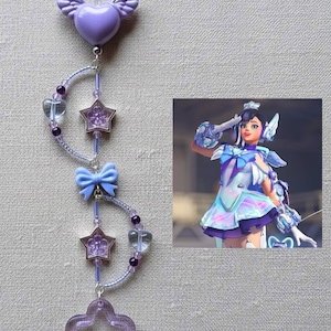 May include: A purple charm with a winged heart, stars, a bow, and heart-shaped beads. The charm is connected by a beaded chain. A digital illustration of a character in a blue and purple outfit is in the background.