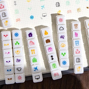 May include: White plastic stamp sets with colorful icons, including coffee cups, cameras, and weather symbols, are arranged on a dotted notebook. A small calendar is visible. The stamps are designed for bullet journaling and planning.