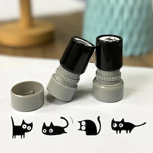 Op de afbeelding: Twee zwarte en grijze stempels met kattenmotief en een bijpassende inktkussendoos. De stempels liggen naast een vel papier met vier verschillende zwarte kattenillustraties. Een lamp en een vaas staan op de achtergrond.