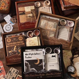 Puede incluir: Cuatro cajas de regalo rectangulares que contienen juegos de papelería. Cada caja incluye un cuaderno, papel decorativo y dos rollos de cinta washi. Las cajas presentan gráficos de estilo vintage, incluyendo un vaquero, una luna creciente y el texto Mist Night Ashen Feather.