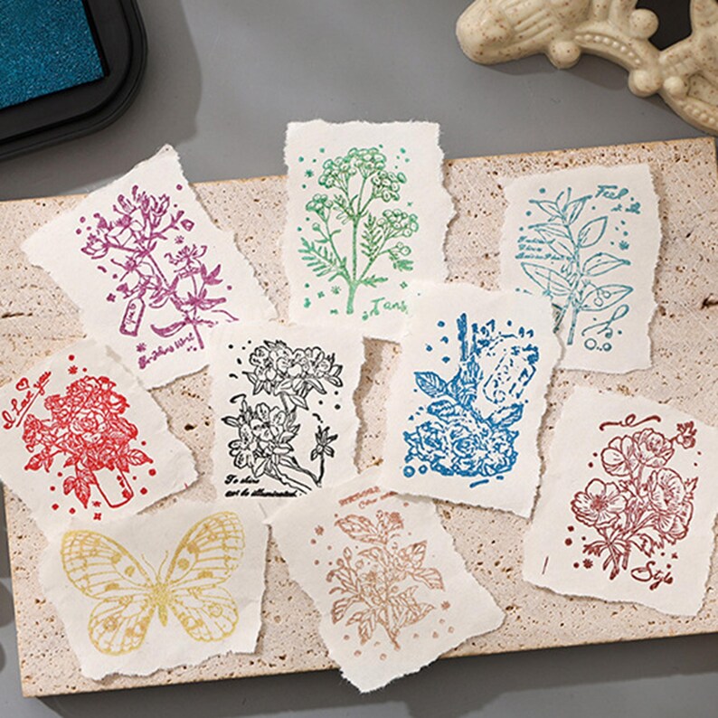 May include: A collection of handmade paper cards with floral and butterfly designs in various colors. The cards feature stamped illustrations of flowers, leaves, and a butterfly. Some cards have text. The cards are arranged on a textured surface.