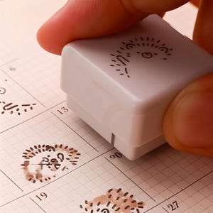 Puede incluir: Un sello blanco con un diseño de sol caprichoso se presiona sobre una página de calendario. El sello tiene una forma pequeña y cuadrada. La página del calendario tiene un patrón de cuadrícula con números impresos y otras imágenes selladas.