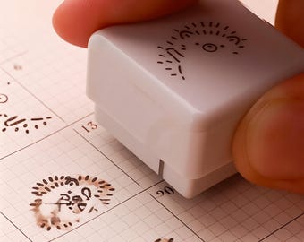 May include: A white stamp with a whimsical sun design is being pressed onto a calendar page. The stamp has a small, square shape. The calendar page has a grid pattern with printed numbers and other stamped images.