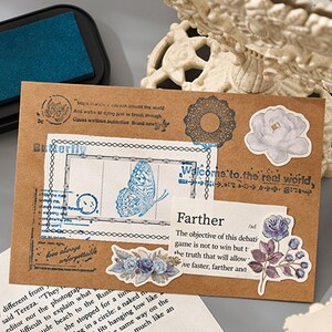 May include: A brown paper envelope with various decorative elements. It features a blue butterfly stamp, floral stickers, and text that reads "Welcome to the real world." The design has a vintage, scrapbook aesthetic.