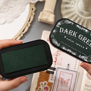 May include: Two black ink pads with dark green ink. One pad is open, revealing the ink surface. The other pad is closed, with the words "DARK GREEN" and "color space" printed on the lid. The ink is water-based.