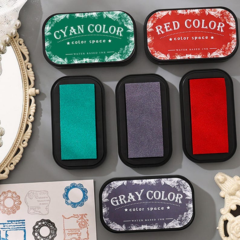 May include: A collection of ink pads in various colors, including cyan, red, and gray, with the text "CYAN COLOR," "RED COLOR," and "GRAY COLOR" on the lids. Each pad is rectangular with rounded corners and a black base. The pads are labeled "color space" and "water based ink."