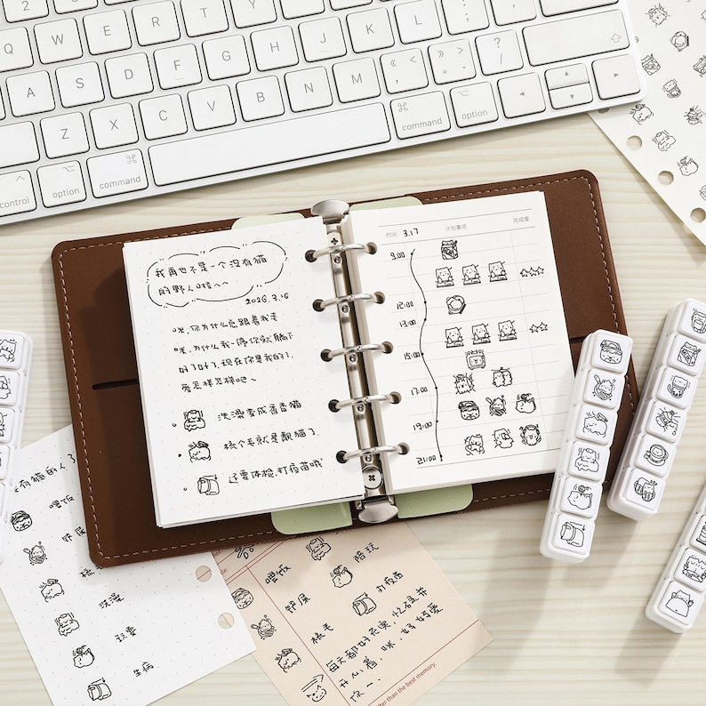 May include: A brown leather six-ring binder planner open to a page with handwritten Chinese text and a daily schedule. The planner features a metal ring mechanism, accompanied by several white plastic stamp blocks and loose paper sheets with small black ink illustrations.
