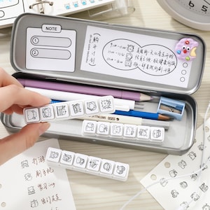 May include: A rectangular metal pencil case containing a purple pencil, a blue pencil, a metallic pen, and a small blue pencil sharpener. Inside the lid is a white note card with text and a purple cookie-shaped magnet. Three white plastic stamp strips with black line art illustrations of cats are displayed.