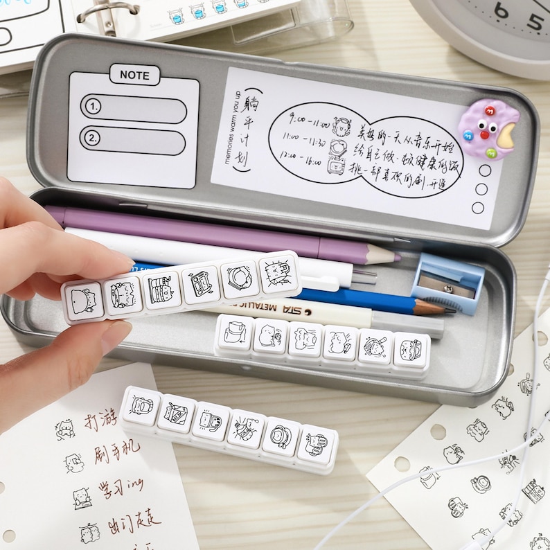 May include: A rectangular metal pencil case containing a purple pencil, a blue pencil, a metallic pen, and a small blue pencil sharpener. Inside the lid is a white note card with text and a purple cookie-shaped magnet. Three white plastic stamp strips with black line art illustrations of cats are displayed.