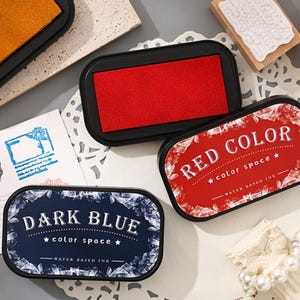 May include: An assortment of ink pads in red and dark blue, with the text "RED COLOR" and "DARK BLUE". The ink pads are water-based and have a rectangular shape with rounded edges. The image also shows a stamp.