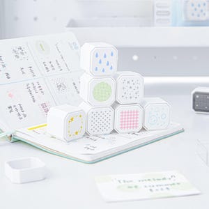 May include: A stack of seven white square rubber stamps with rounded corners, each featuring a unique pattern on the face including blue raindrops, green dots, silver stars, yellow stars, black polka dots, pink gingham, and blue bubbles.