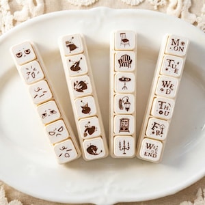 May include: Four sets of decorative stamps on a white plate. Each set has a series of small, square stamps with various designs and text. The stamps feature faces, furniture, and days of the week.