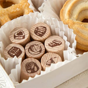 Puede incluir: Una colección de siete sellos de madera cilíndricos, cada uno con una ilustración única en tinta marrón de diferentes dulces de chocolate, dispuestos dentro de una caja forrada de papel blanco junto a dos galletas de azúcar redondas.