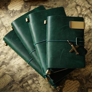 Puede incluir: Un conjunto de diarios de cuero verde oscuro con detalles de latón. Cada diario tiene un cierre elástico a juego y un pequeño dije de avión. Los diarios están colocados sobre un mapa antiguo.