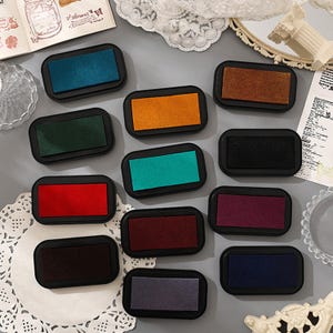 May include: A collection of rectangular ink pads in various colors, including teal, orange, green, red, and purple. Each pad has a black plastic casing. The pads are arranged on a light-colored surface, suggesting a craft or stationery theme.