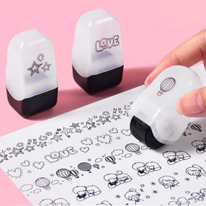 Könnte beinhalten: Weißer und schwarzer Rollstempel, der ein dekoratives Design erzeugt. Der Stempel rollt über weißes Papier und erzeugt ein Muster aus Sternen, Herzen, Heißluftballons und Paaren. Zwei weitere Stempel sind sichtbar, einer mit Sternen und der andere mit dem Wort "LOVE".