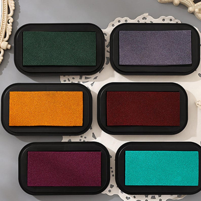May include: A collection of six ink pads in various colors, including green, purple, orange, burgundy, dark purple, and teal. Each ink pad is rectangular with rounded corners and set in a black plastic tray. The pads are arranged on a white, lace-edged surface.