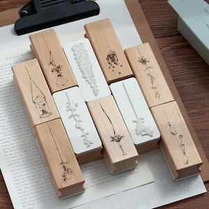 May include: A collection of eight rectangular rubber stamps with light-coloured wooden handles. Each stamp features a unique, delicate black line drawing of botanical elements, including hanging plants, flowers, and vases, printed on the wood surface.