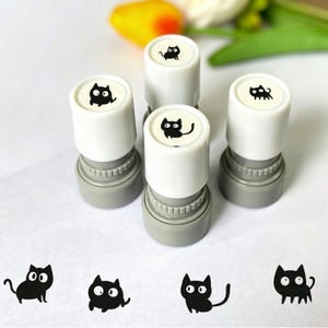 May include: Four cylindrical self-inking stamps with white tops and grey bases. Each top features a unique black and white cartoon cat illustration. Four corresponding black ink cat impressions are stamped on the surface below the stamps.