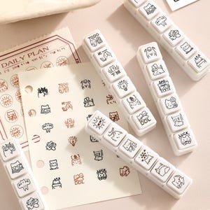 May include: White plastic stampers featuring black cat illustrations and text including "TODO" and "DDL". The image also shows a "DAILY PLAN" sheet and cat-themed stickers, all in a minimalist style.