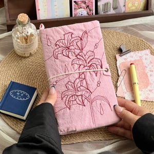May include: A rectangular notebook with a textured light pink fabric cover featuring dark red embroidered lily flowers and thin line art details. The notebook is secured with a thin white elastic band wrapped horizontally around the center.