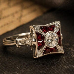 Può includere: Un anello d'argento con un elemento centrale quadrato, rosso rubino e tempestato di diamanti. Il centro presenta un grande diamante rotondo e trasparente. Il gambo dell'anello è anch'esso ornato da piccoli diamanti, creando un look vintage ed elegante.