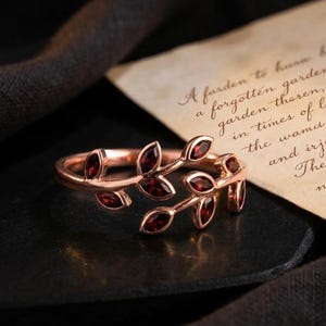 May include: Rose gold ring with a leaf design. The ring has small, oval-shaped, red gemstones set along the leaves. The ring is displayed on a dark surface with a handwritten note in the background.