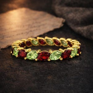 May include: A gold-toned ring featuring alternating red and green oval-cut gemstones. The gemstones are set closely together, creating a continuous band of color. The ring is displayed on a dark, textured surface.